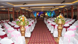 A banquet hall with rows of tables, white and red chair covers, and golden vases with flowers. - Hotel Basant Residency (Bengaluru)