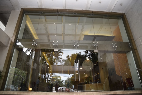 A modern building entrance with glass doors, a visible push button, and a reflection of palm trees and a car outside.
