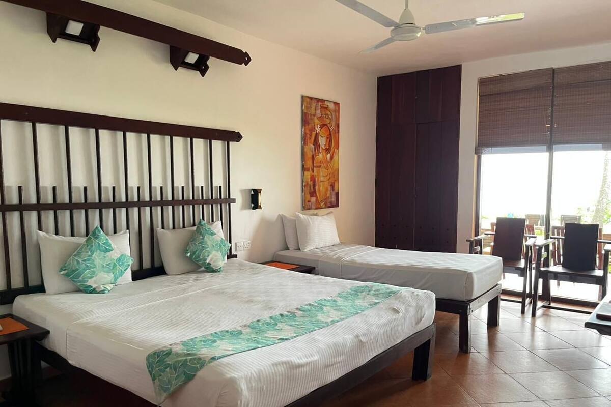 A hotel room with two beds, a ceiling fan, a painting, and a view of the outdoors.