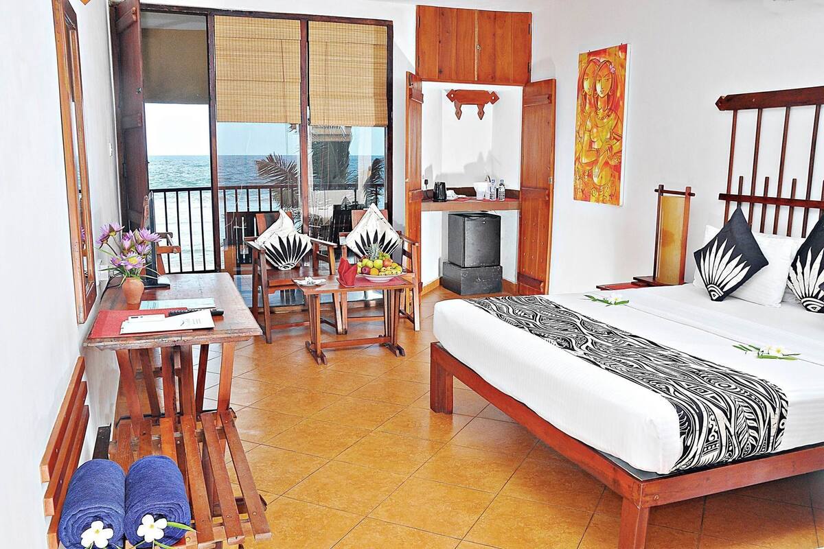 A hotel room with a bed, a dining table with chairs, a kitchenette, and a balcony with a view of the sea.