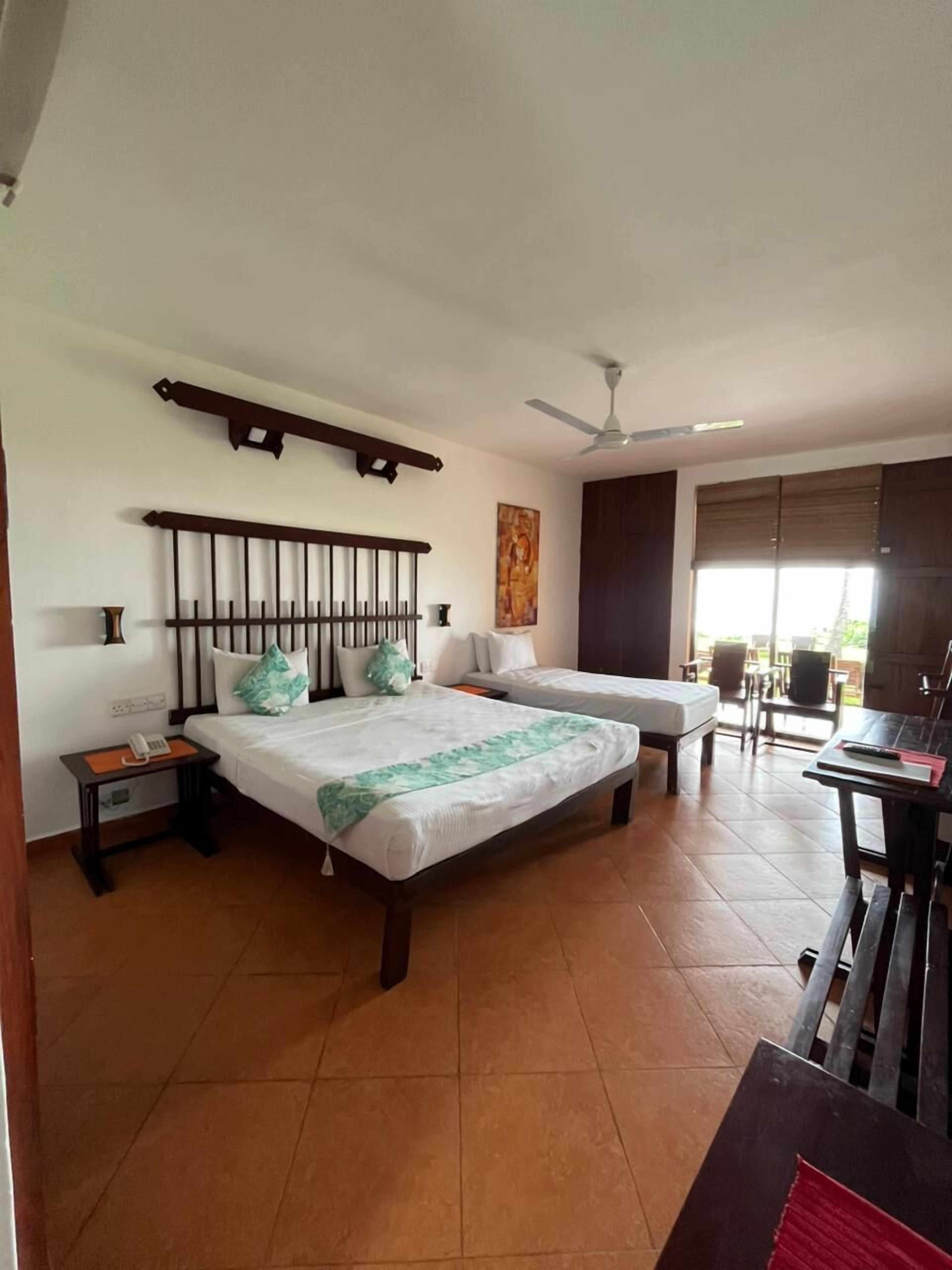 A hotel room with two beds, a ceiling fan, a painting, and a window with blinds.