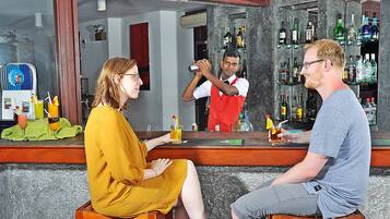 Zona de bar con un camarero preparando bebidas, una mujer sentada y un hombre sentado frente a ella.