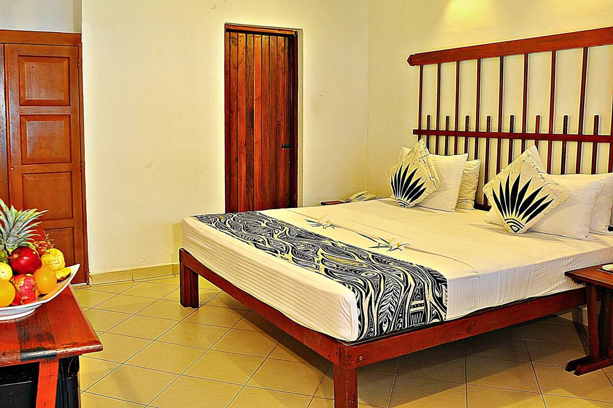 A hotel room with a wooden bed, a bedside table with a fruit bowl, and a wooden door.
