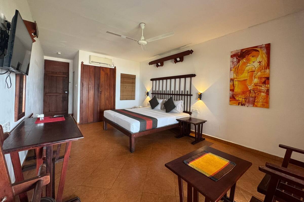 A hotel room with a bed, a desk, a chair, a TV, a ceiling fan, and a painting on the wall.