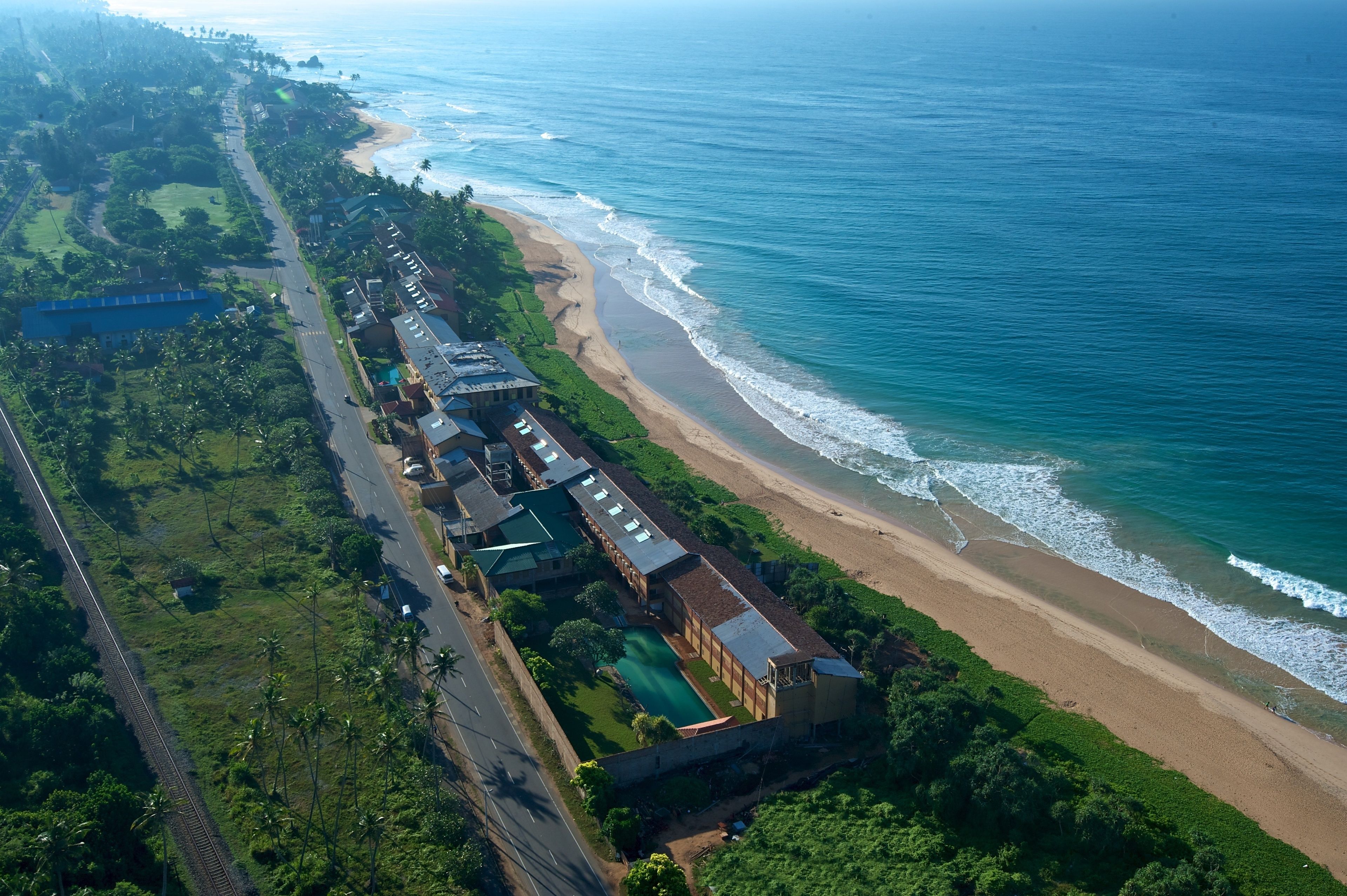 An aerial view of a coastal area with a beach, a road, and buildings with a pool.