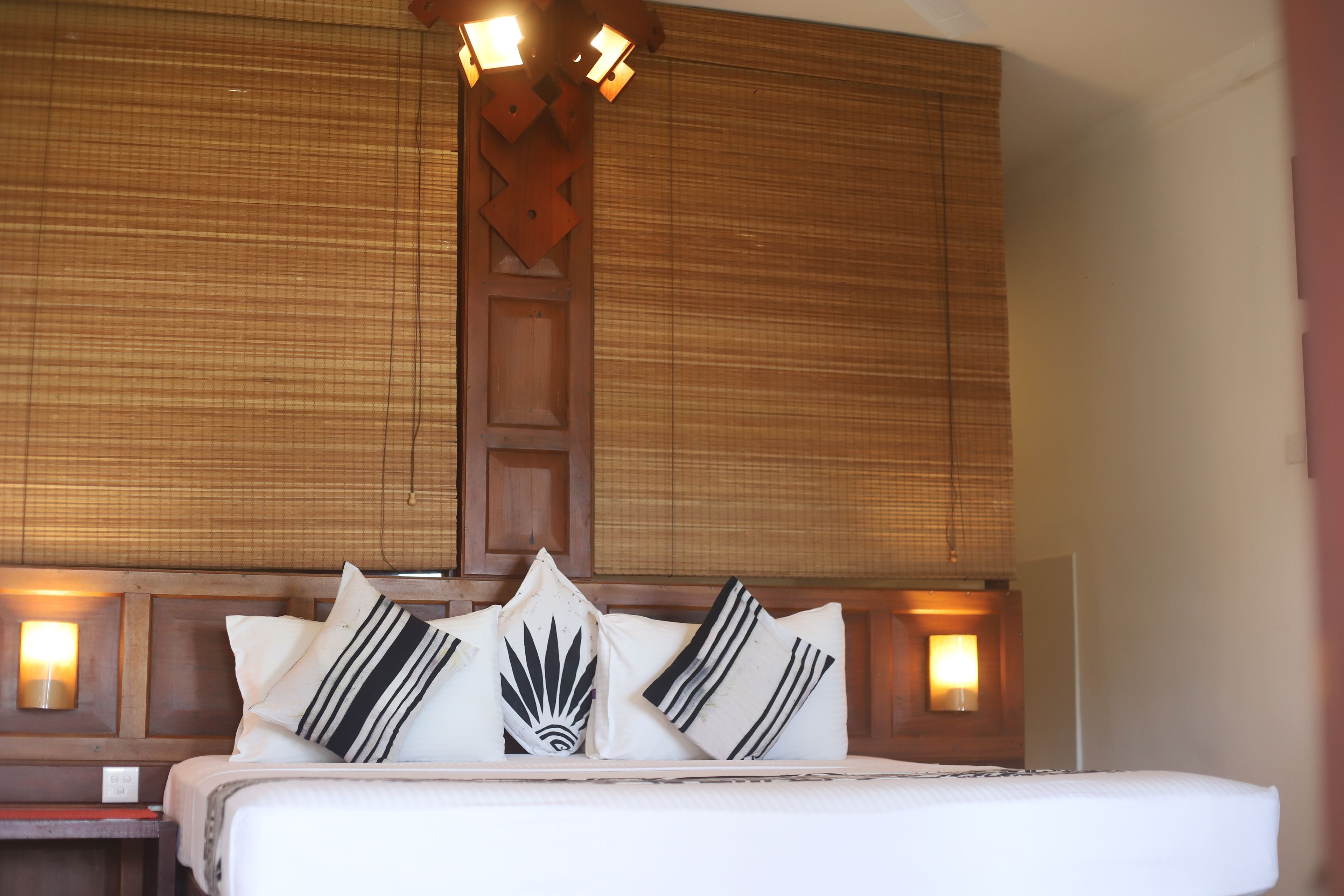 Una habitación de hotel con una cama grande, varios almohadones, un cabecero de madera y lámparas fijadas a la pared.