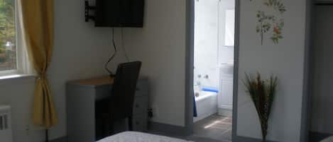 A bedroom with a bed, a desk with a chair, a TV mounted on the wall, a bathroom visible through an open door, and a decorative plant.