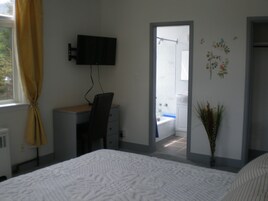 A bedroom with a bed, a desk with a chair, a TV mounted on the wall, a bathroom visible through an open door, and a decorative plant.