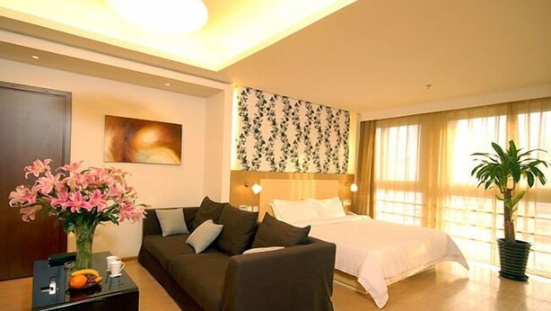 Un cuarto de hotel con una cama, un sofá, una mesita de noche con un jarrón de flores, un cuadro en la pared y un ventanal con cortinas.