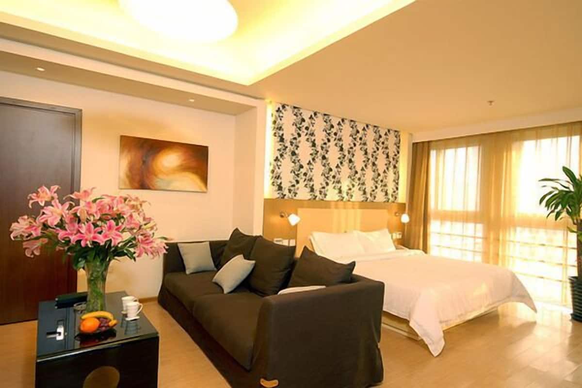A hotel room with a bed, a sofa, a nightstand with a vase of flowers, a painting on the wall, and a large window with curtains.
