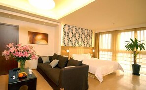 Un cuarto de hotel con una cama, un sofá, una mesita de noche con un jarrón de flores, un cuadro en la pared y un ventanal con cortinas.