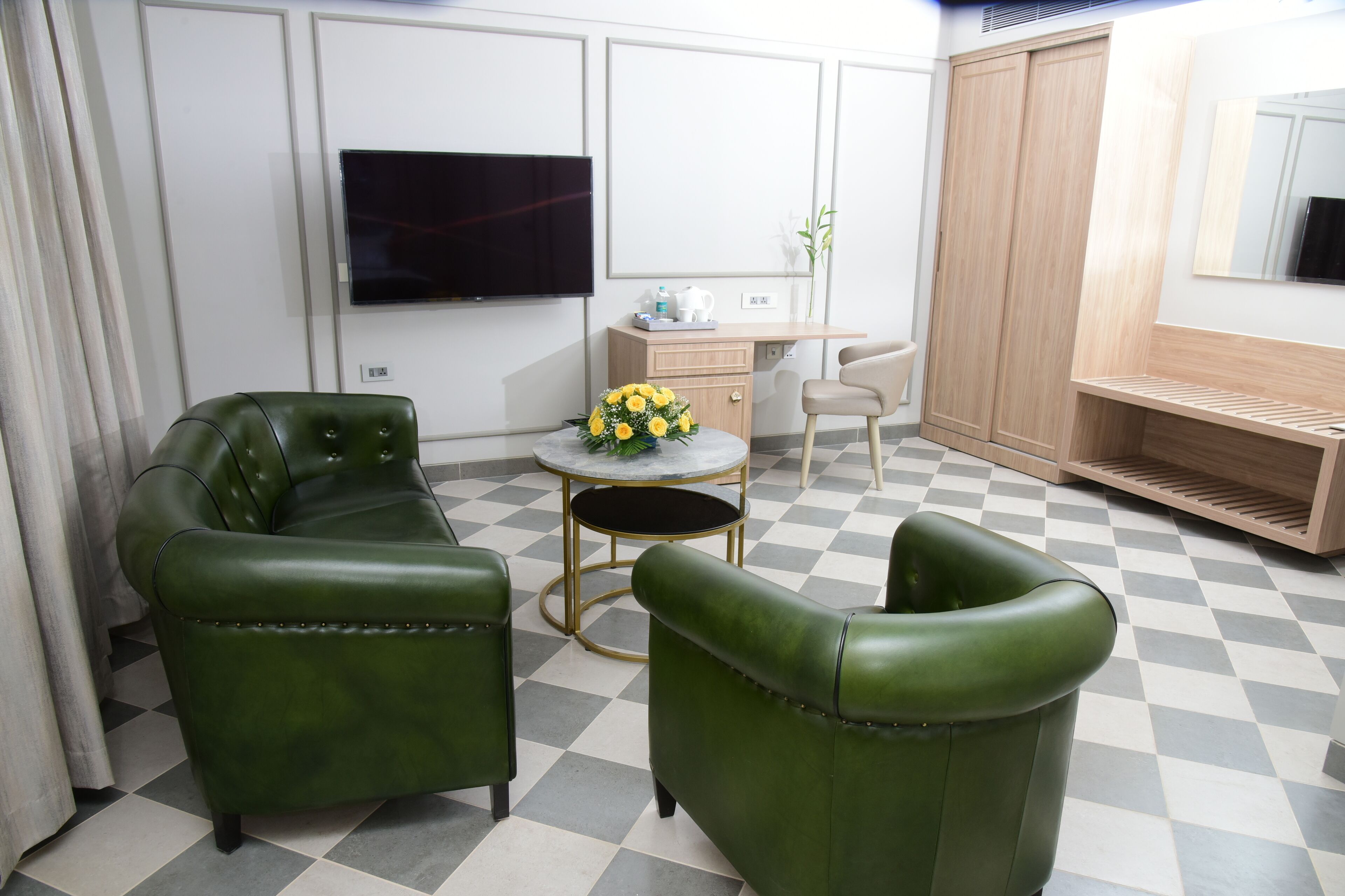 A room with two green leather sofas, a small round table with a yellow flower arrangement, a wooden desk, a television mounted on the wall, and a wardrobe.