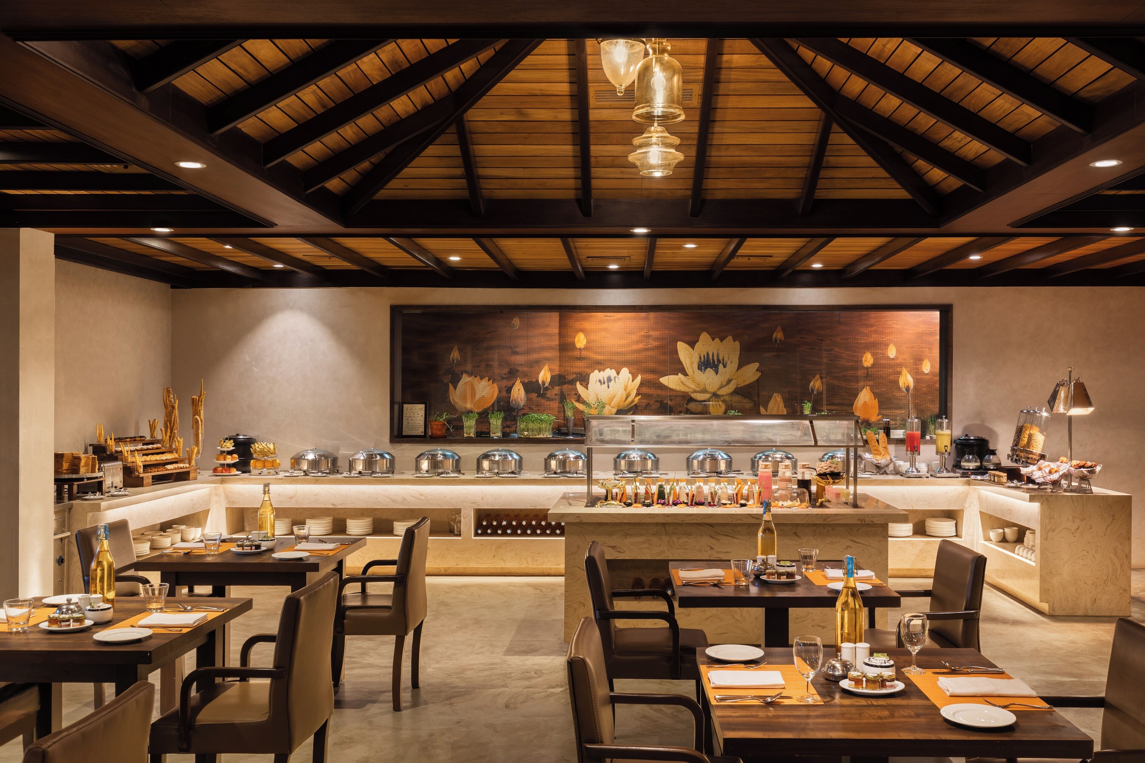 Restaurant amb un sostre alt, biges de fusta i un mural floral gran. Taules preparades amb plats, gots i coberts.
