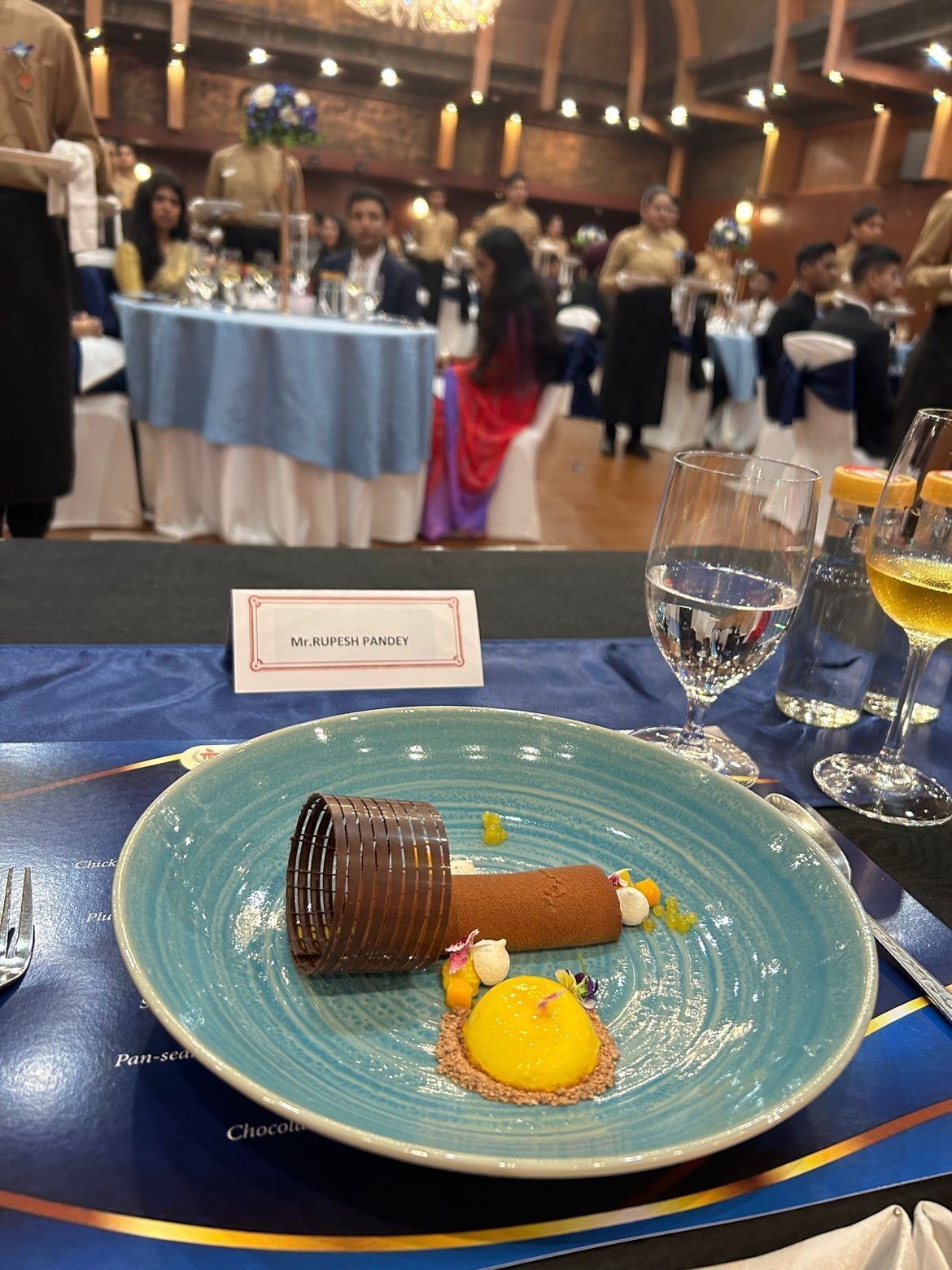 Un piatto dessert con un cilindro di cioccolato, una sfera gialla e piccoli decori su un piatto blu, con un cartellino che riporta il nome 