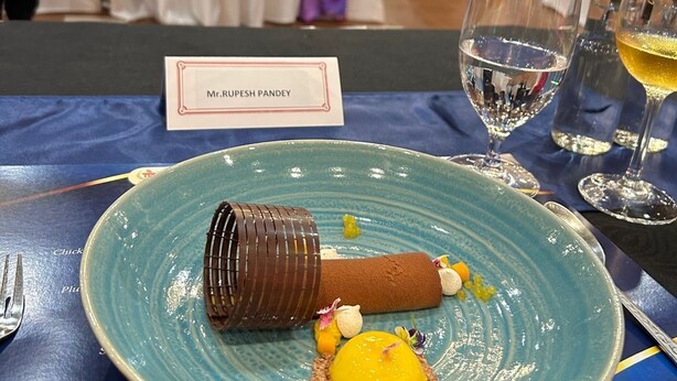 A dessert plate with a chocolate cylinder, a yellow sphere, and small garnishes on a blue plate, with a name card reading "Mr. Rupesh Pandey" in the background. - Fortune Valley View, Manipal - Member ITC Hotels' Group (Udupi)