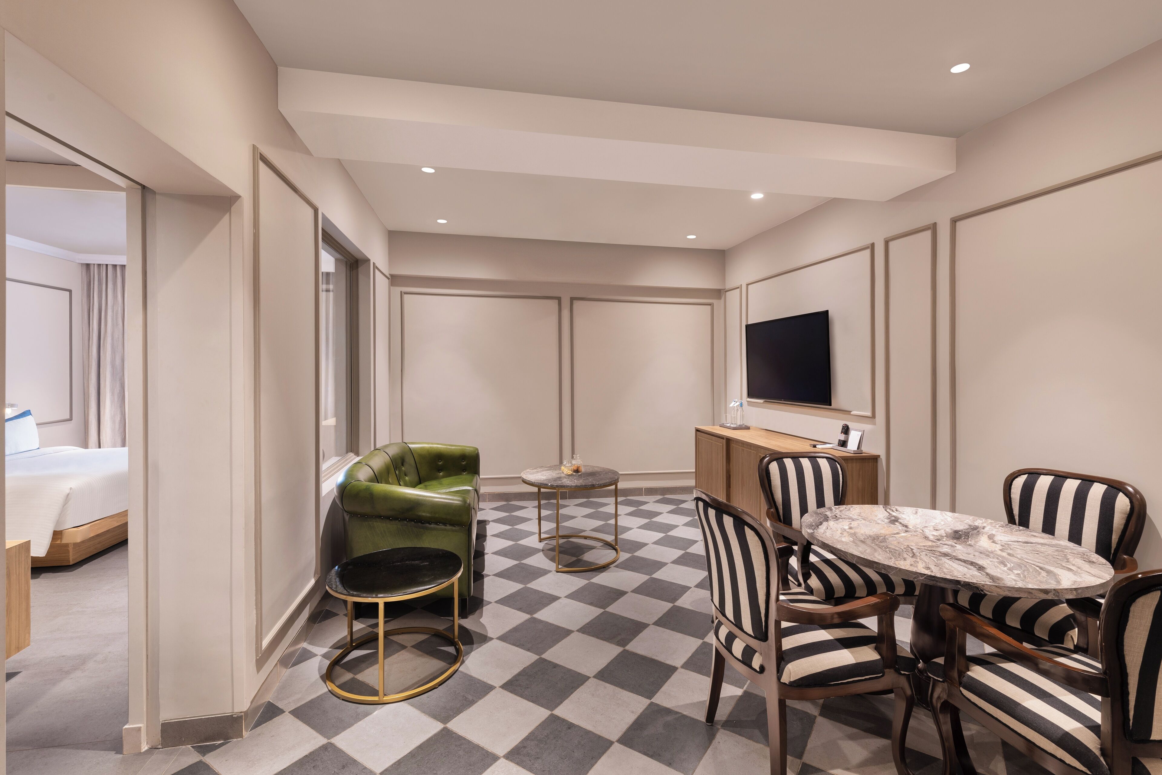 Habitación de hotel con suelo a cuadros, un sillón verde, sillas a rayas, una mesa redonda y un televisor en la pared.