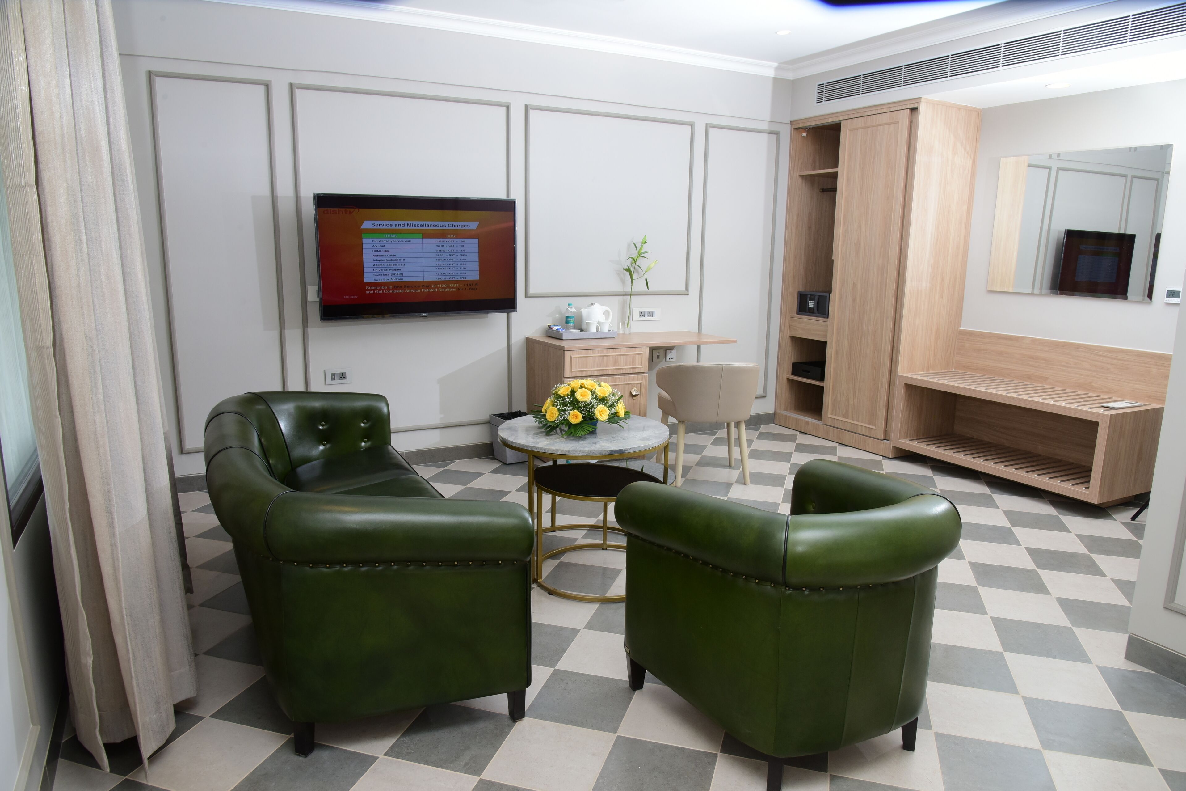 Une chambre d’hôtel avec un sol à carreaux, deux fauteuils en cuir vert, une petite table ronde ornée d’un vase de fleurs jaunes, un téléviseur fixé au mur et une armoire en bois.