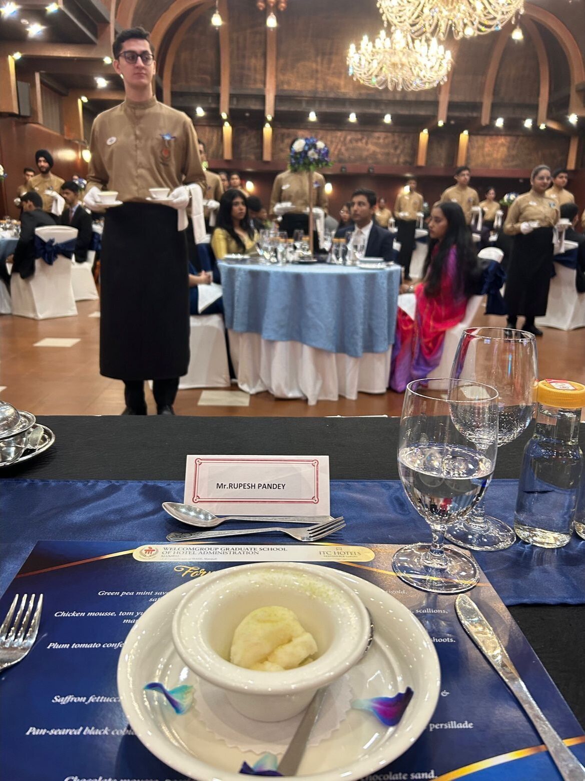 A formal dining setting with a waiter, a table with a name card reading "Mr. RUPESH PANDEY", and a menu with various dishes.