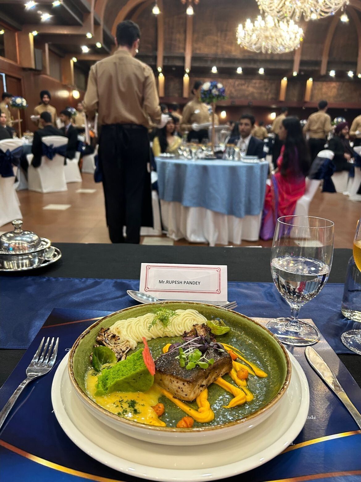 Un ambiente formal para cenar con un plato de comida, una tarjeta de nombre y un vaso de agua sobre una mesa.