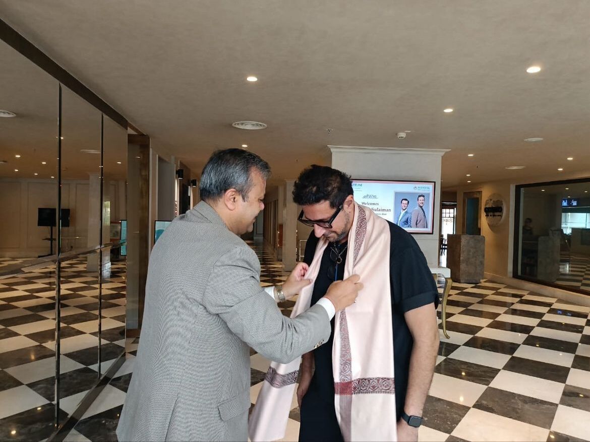 Two men in a hotel lobby with a checkered floor and a projection screen in the background.