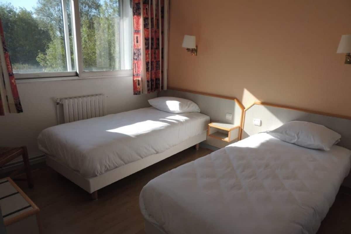 A hotel room with two single beds, a wooden nightstand, a chair, and a window with curtains.