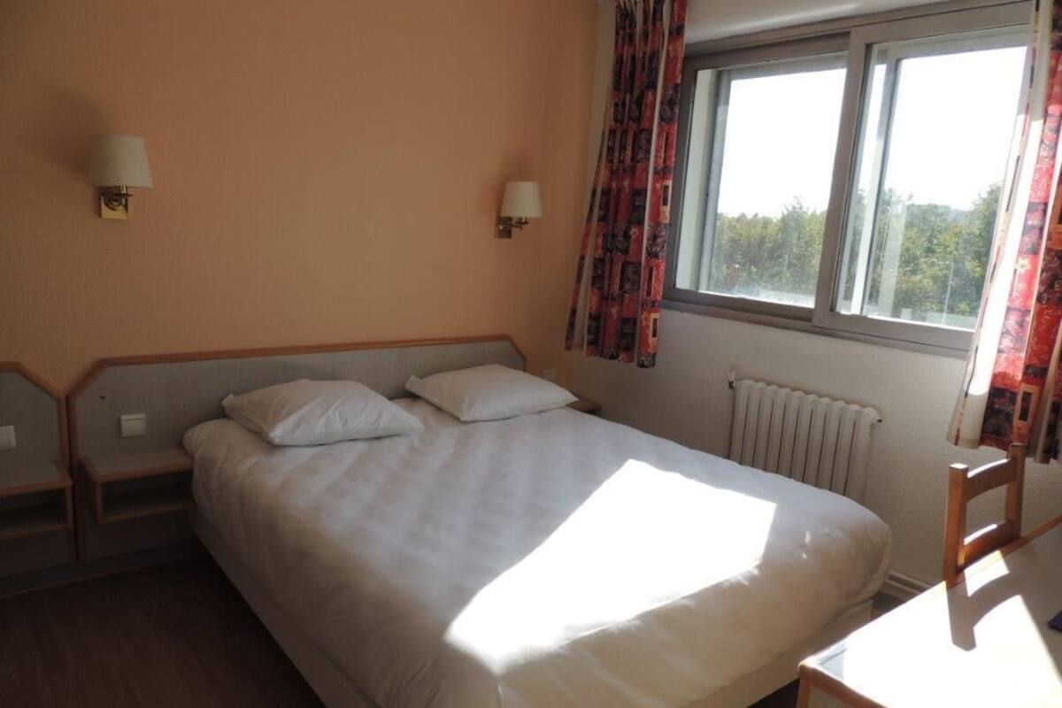 A hotel room with a single bed, two bedside tables, a chair, a window with curtains, and a radiator.