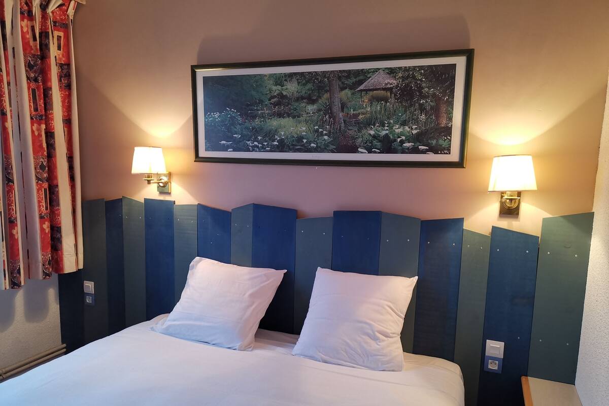 A hotel room with a large bed, two bedside lamps, a framed nature picture on the wall, and a window with curtains.