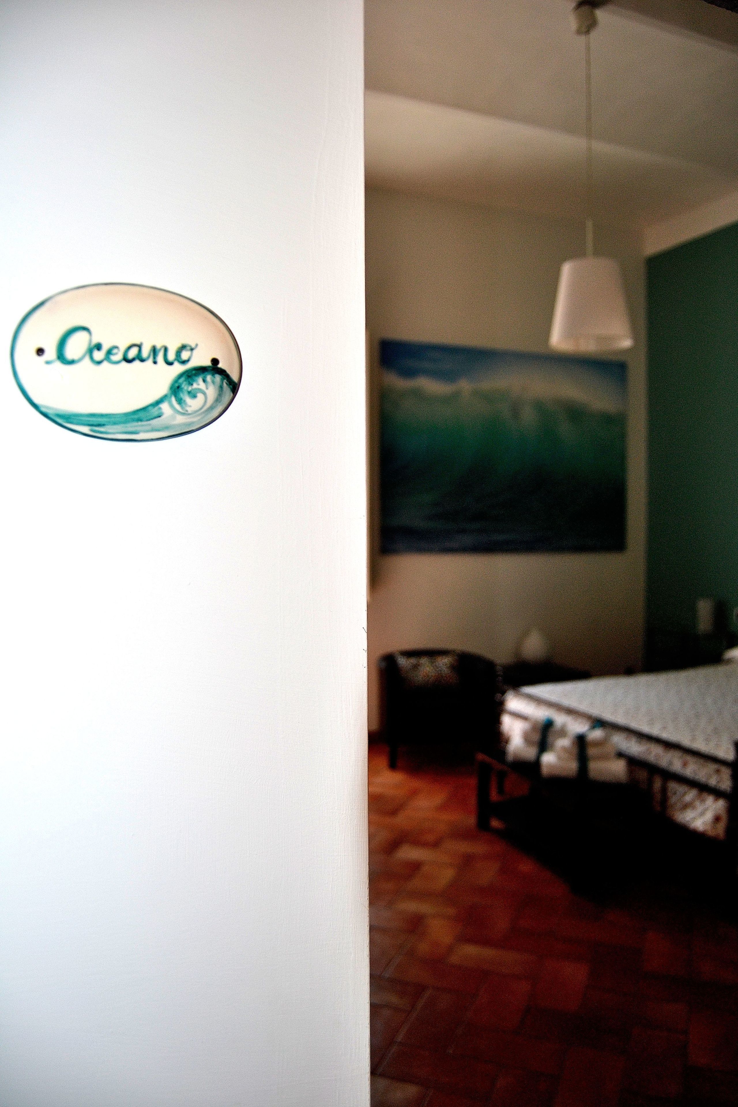 A room with a bed, a chair, a painting of a landscape, and a logo on the wall.