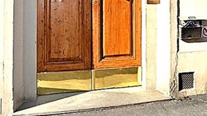 Une porte à deux battants en bois, dotée d’une poignée et d’un heurtoir en laiton, est encastrée dans un mur blanc, au-dessus duquel on aperçoit un élément décoratif en métal noir.