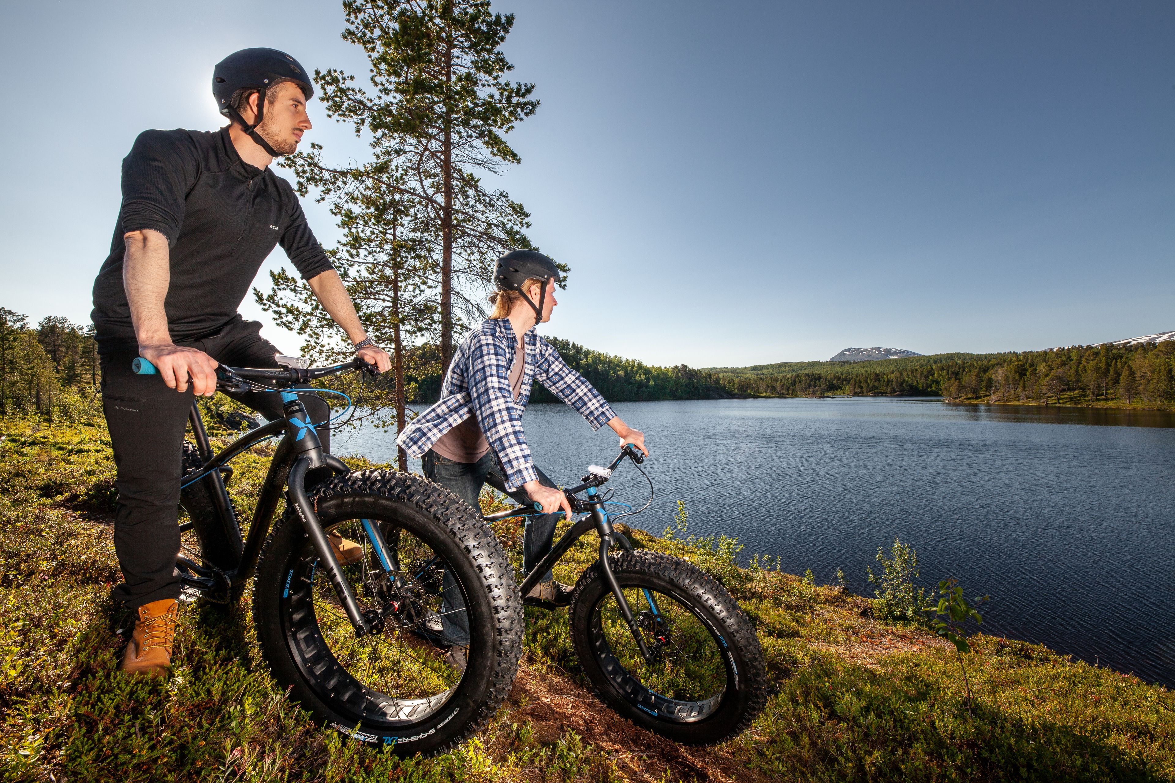 Two people on bicycles by a lake with a forest in the background.