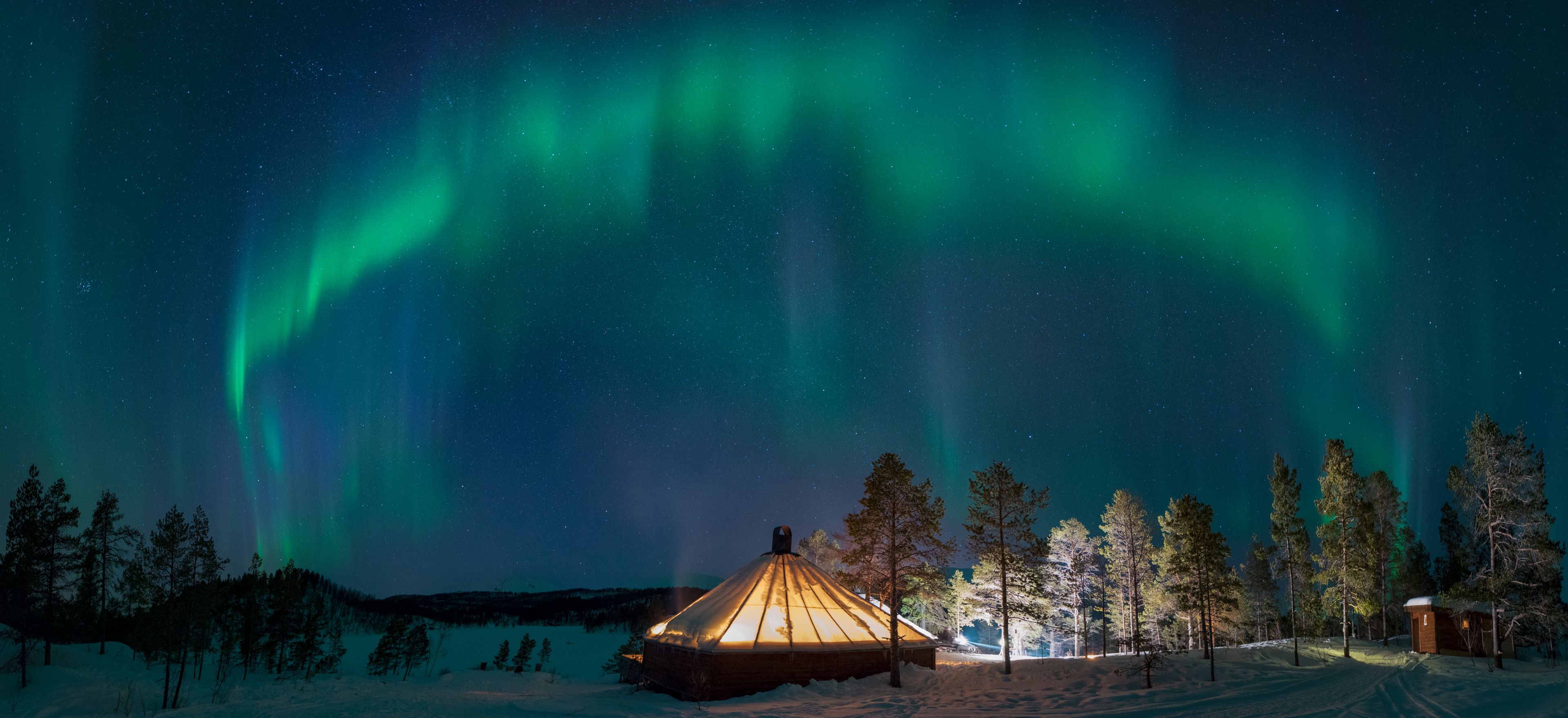 Un paisaje nevado con una yurta de madera iluminada por luces cálidas, rodeada de árboles cubiertos de nieve y un cielo nocturno despejado.