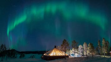 Un paisaje nevado con una yurta de madera iluminada por luces cálidas, rodeada de árboles cubiertos de nieve y un cielo nocturno despejado.