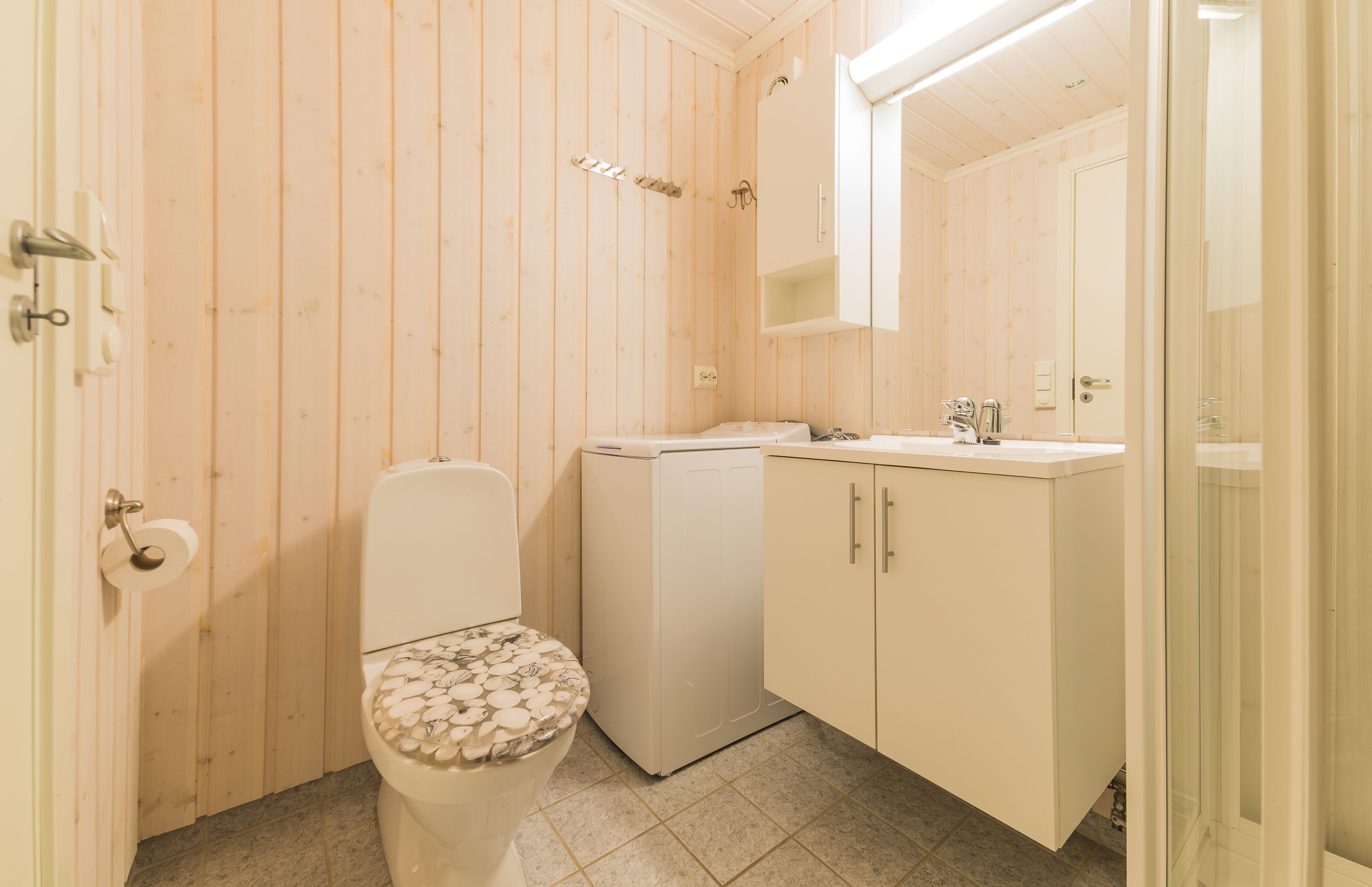 Un baño pequeño y funcional con paredes de madera, un inodoro con asiento decorativo, una lavadora y un lavamanos con espejo.