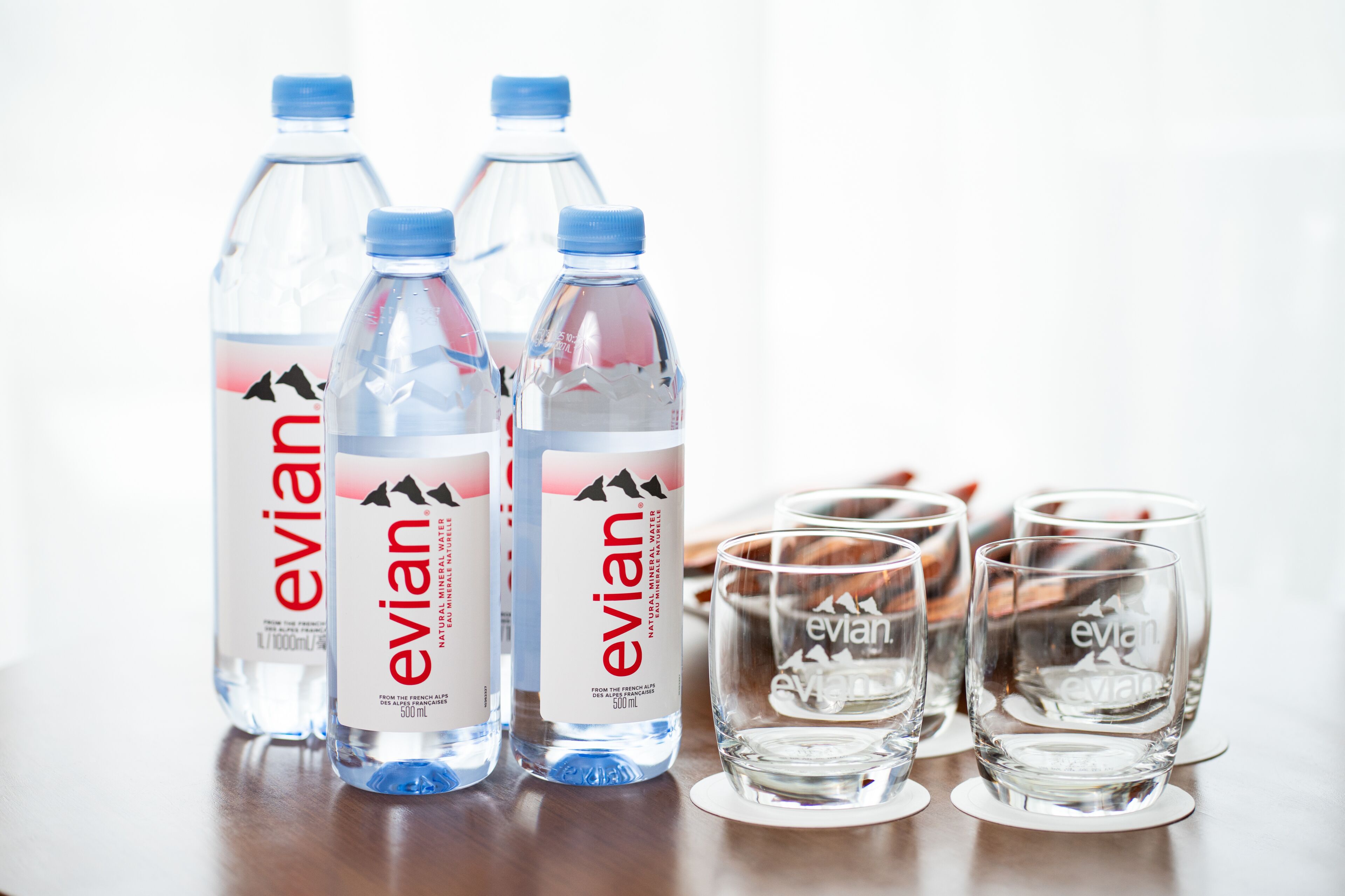 Three Evian water bottles and four glasses on a wooden surface.