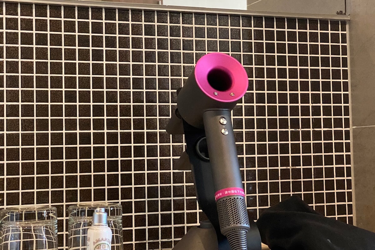 A hair dryer on a counter with a tiled backsplash, a bottle of soap, and a black bag.