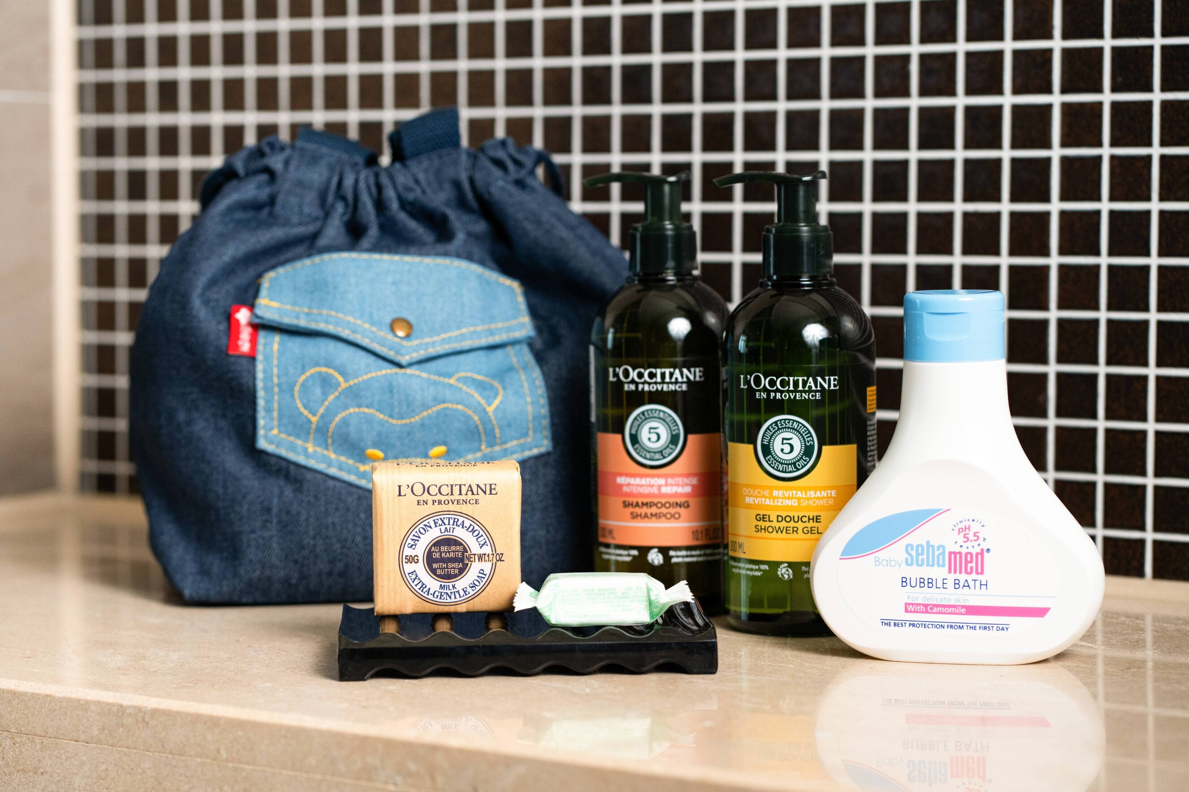 A bathroom counter with a blue drawstring bag, two bottles of L'OCCITANE shampoo, a bottle of Baby Sebamed bubble bath, and a bar of soap.
