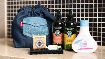 A bathroom counter with a blue drawstring bag, two bottles of L'OCCITANE shampoo, a bottle of Baby Sebamed bubble bath, and a bar of soap.