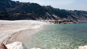 Una playa costera con aguas cristalinas, un litoral rocoso y un cerro cubierto de bosque al fondo.