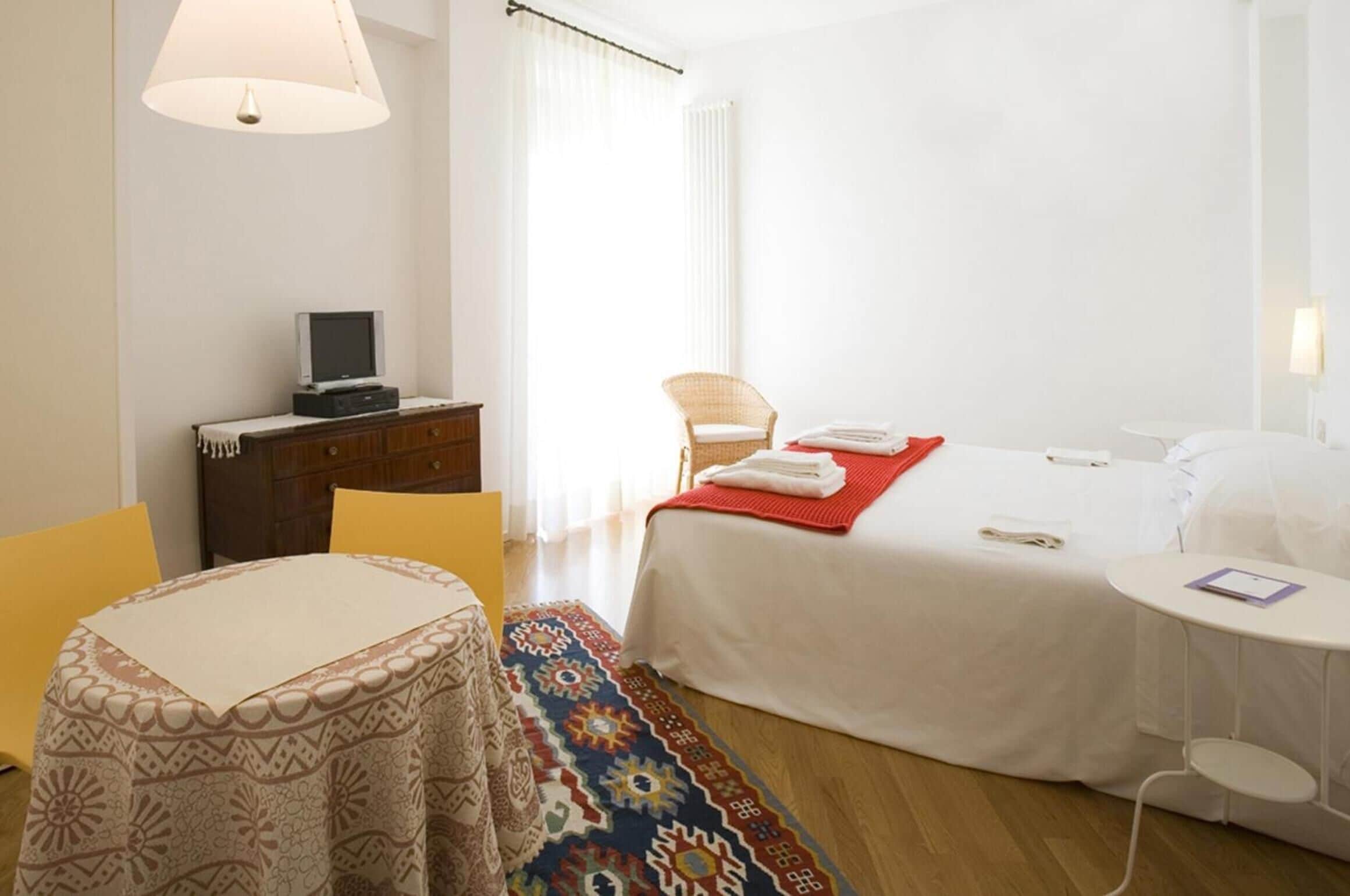 A hotel room with a bed, a small table, a chair, a desk with a television, and a colorful rug.