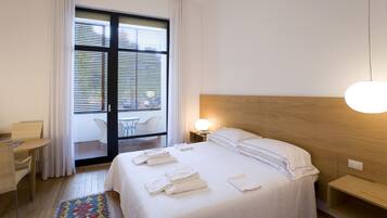 A hotel room with a bed, bedside table, lamp, and a view through a window with blinds.