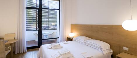 A hotel room with a bed, bedside table, lamp, and a view through a window with blinds.