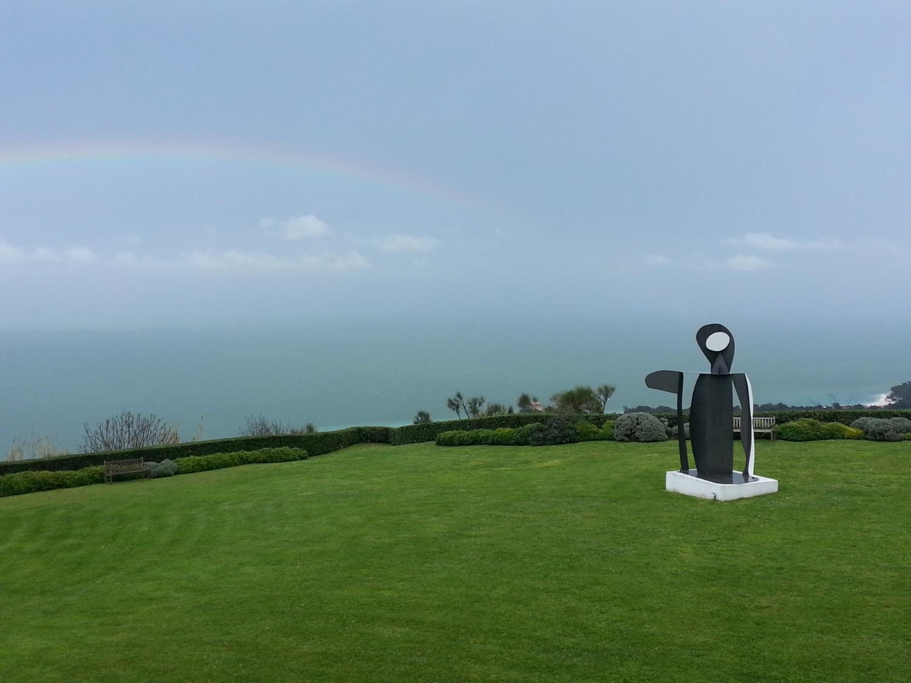 A sculpture on a grassy lawn with a view of the ocean and a rainbow in the background.