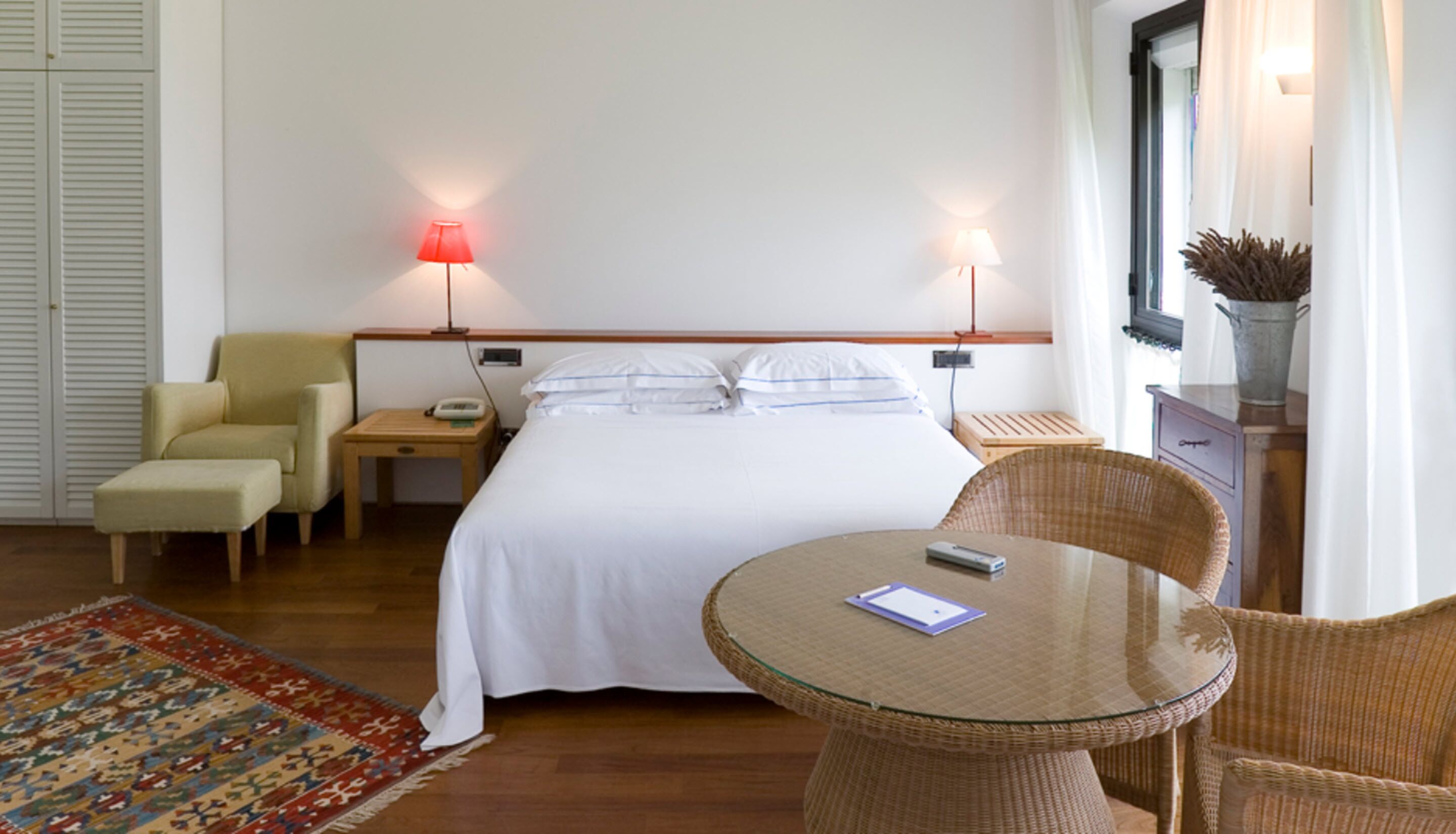 A hotel room with a bed, two bedside lamps, a chair, a small table, a nightstand, and a decorative vase.