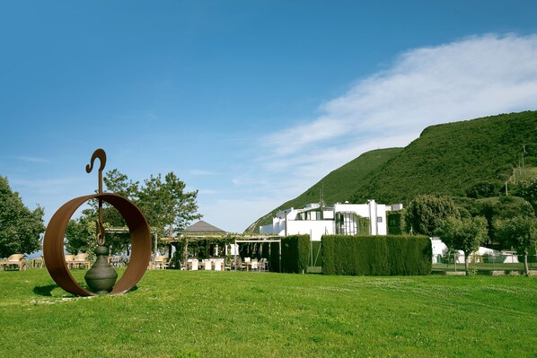 A grassy outdoor area with a unique metal sculpture, a building with a modern design, and a hill in the background.