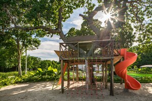 A treehouse playground with a slide, surrounded by greenery and a clear sky.