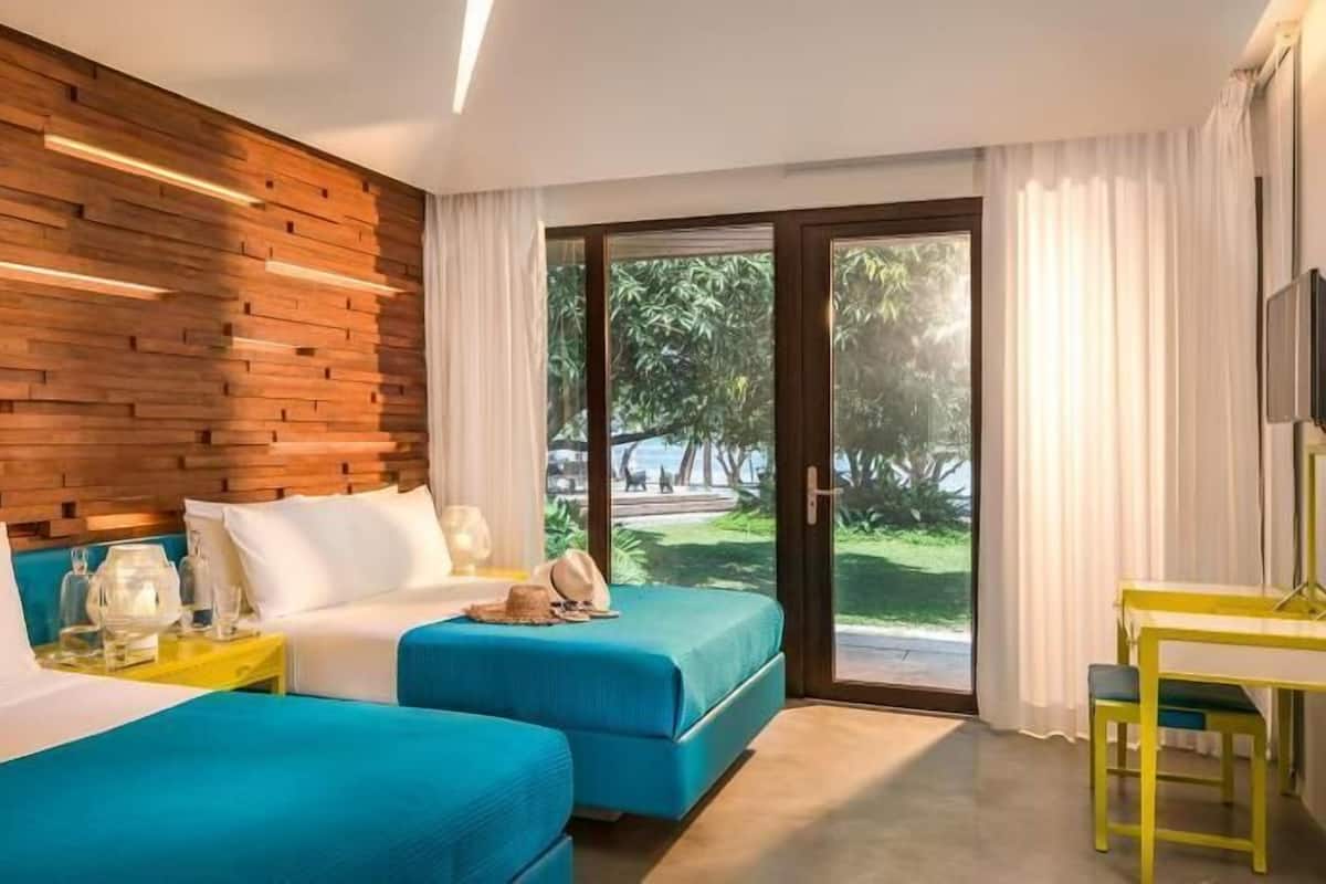 Habitación de hotel con dos camas, un cabecero de madera, un amplio ventanal corredero al exterior y un escritorio amarillo.