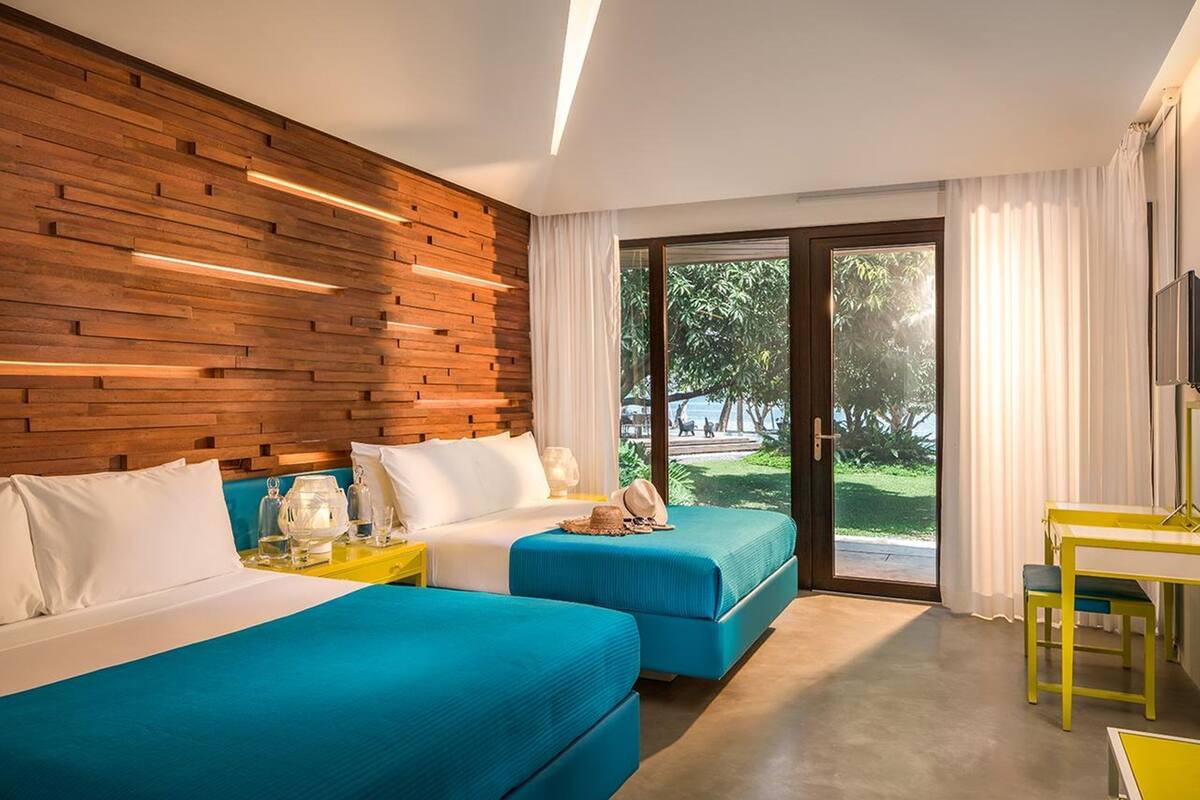 Habitación de hotel con dos camas, un muro decorativo de madera, un amplio ventanal corredero al exterior y un escritorio amarillo.
