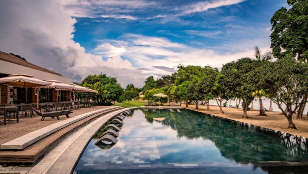 A swimming pool with a tiled deck, surrounded by trees and a building with outdoor seating. - Astoria Palawan (Puerto Princesa)