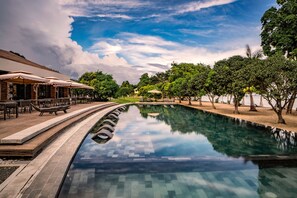 A swimming pool with a tiled deck, surrounded by trees and a building with outdoor seating. - Astoria Palawan (Puerto Princesa)