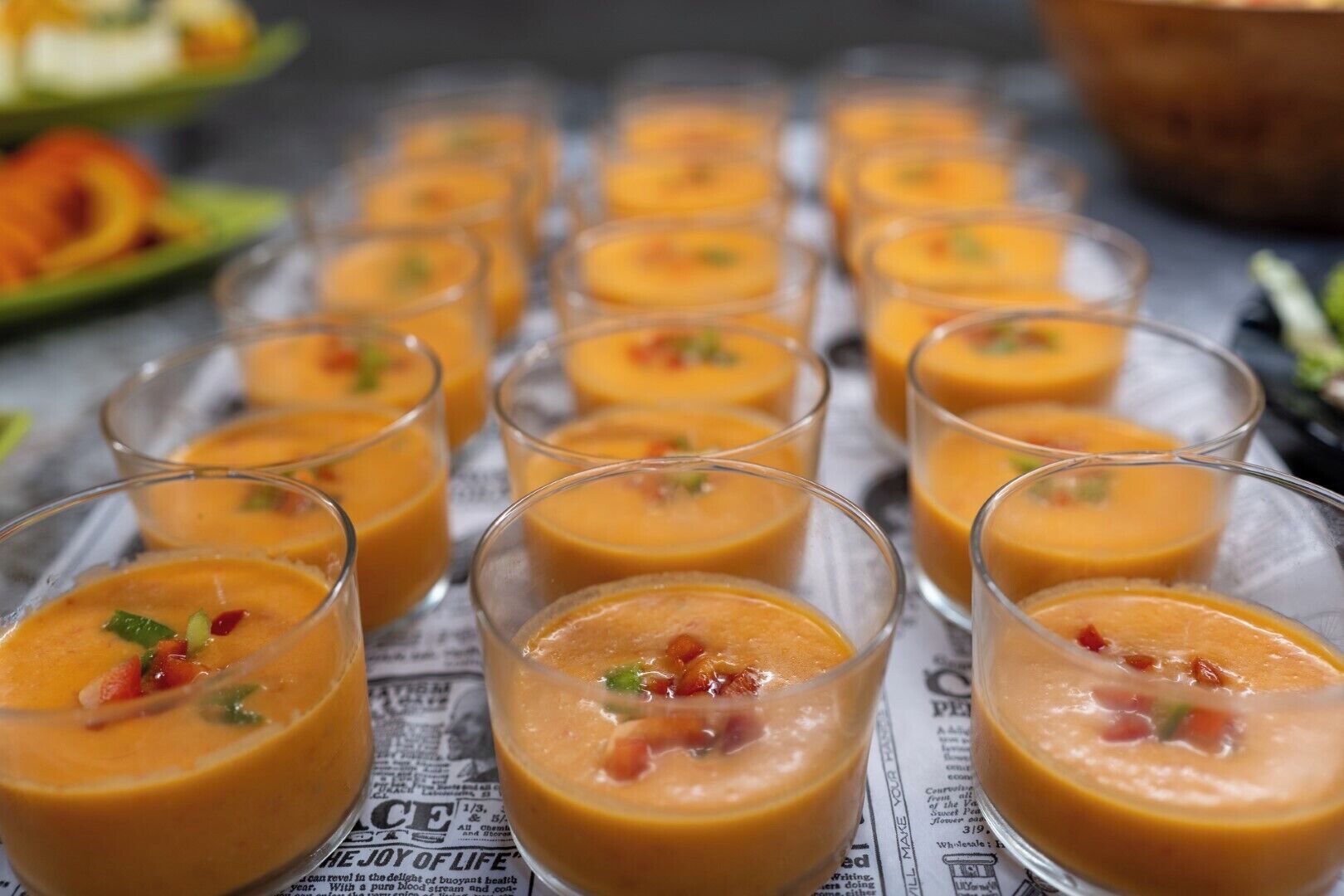 A series of clear glasses filled with a creamy orange soup, garnished with green herbs and red bits, arranged on a surface with newspaper text.